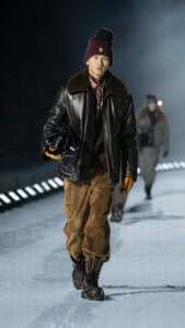 Man in a burgundy knit beanie, leather jacket with fur collar, brown corduroy pants, and gloves walking on an icy runway while holding a black helmet with a small flag.
