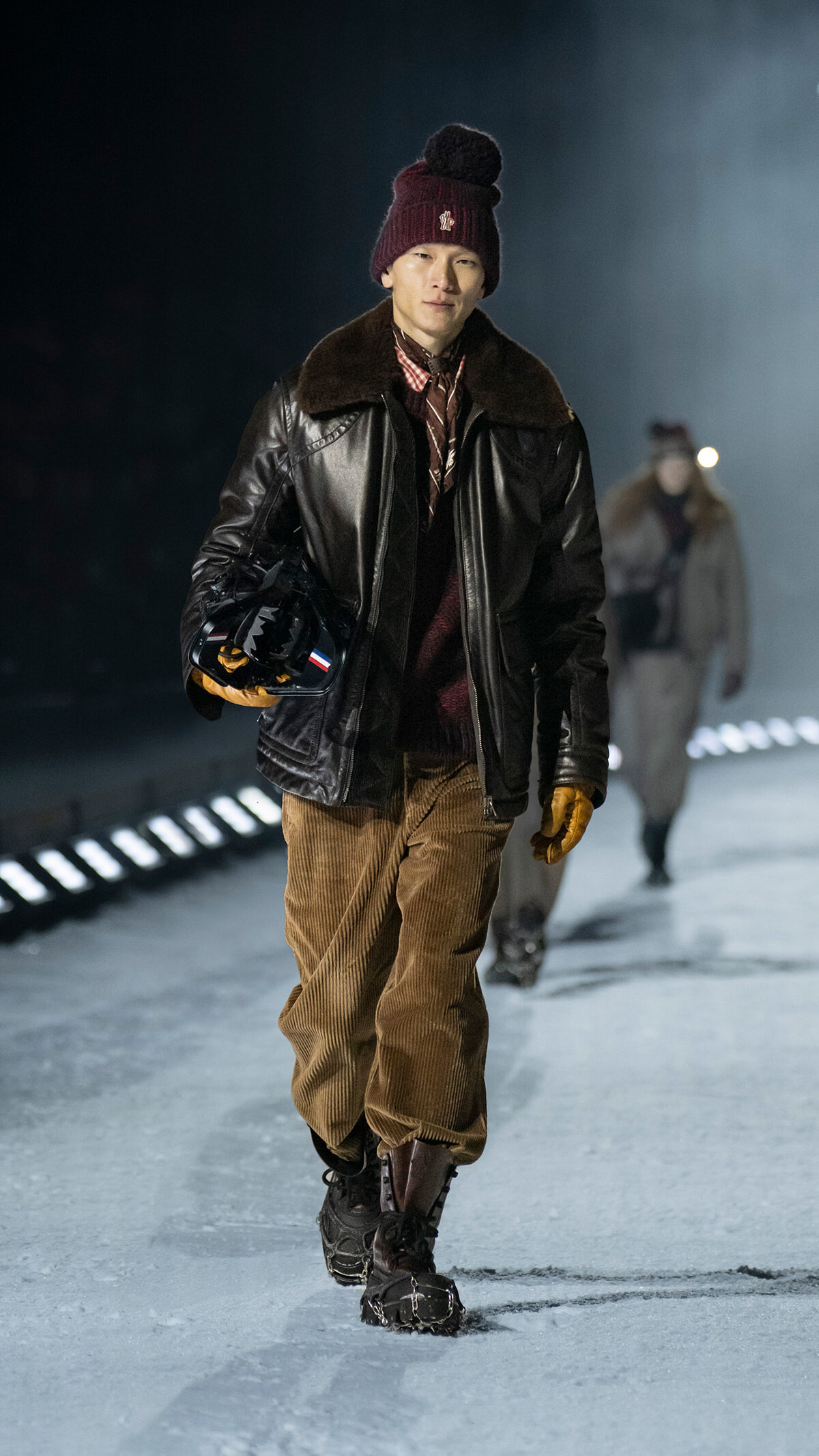 Man in a burgundy knit beanie, leather jacket with fur collar, brown corduroy pants, and gloves walking on an icy runway while holding a black helmet with a small flag.
