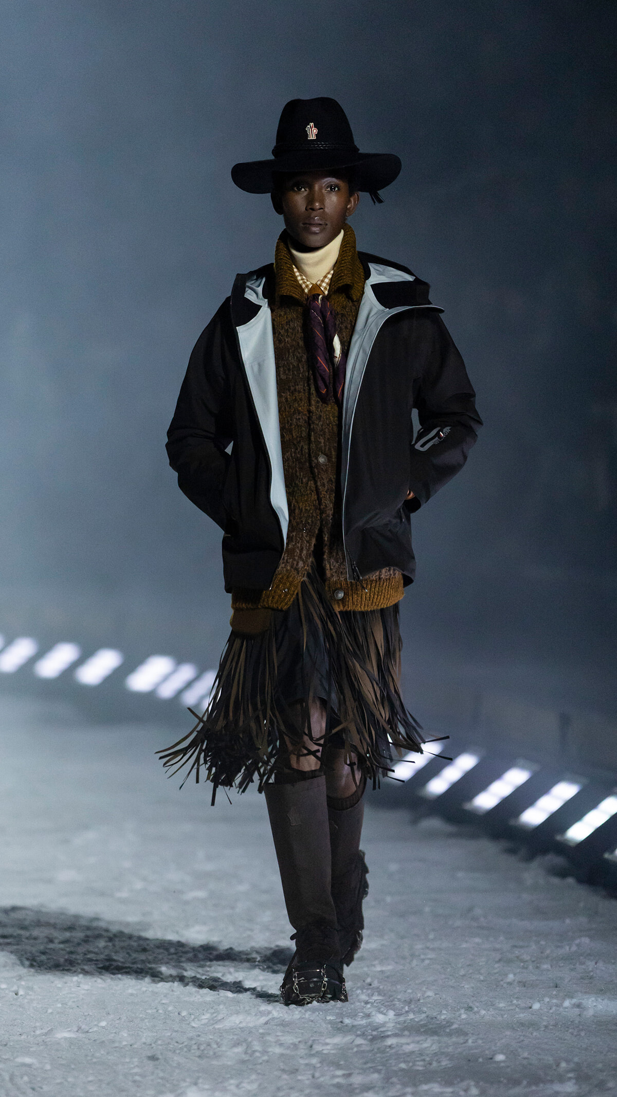 Model walking on a snowy runway in a wide-brim black hat, layered jacket, brown fringe skirt, and tall boots.