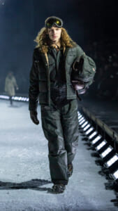 Model walking on a snowy runway wearing a dark winter jacket, pants, gloves, and ski goggles, carrying a helmet under one arm.