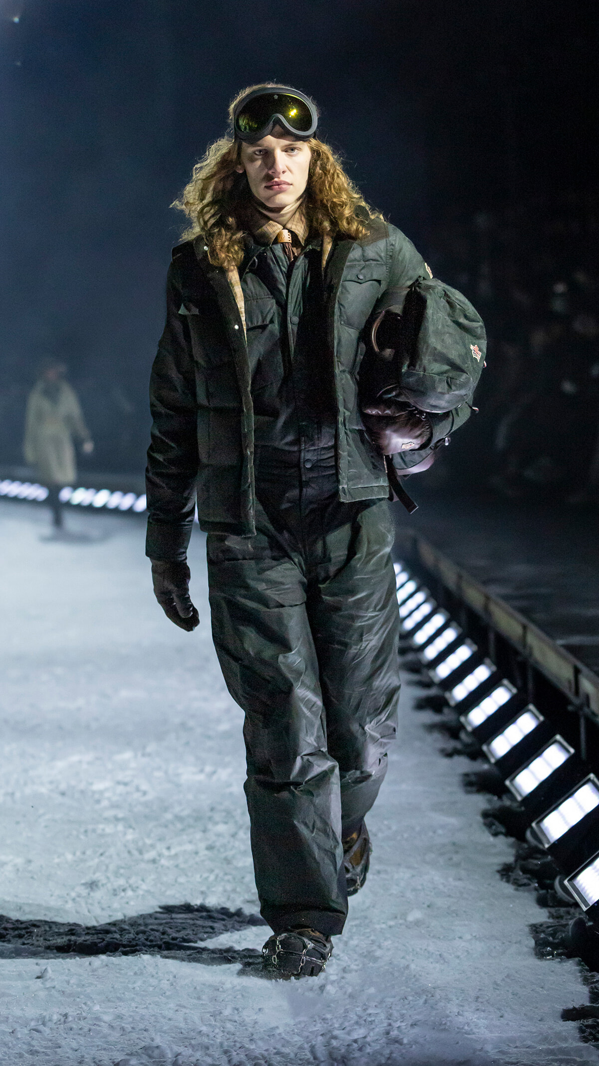 Model walking on a snowy runway wearing a dark winter jacket, pants, gloves, and ski goggles, carrying a helmet under one arm.