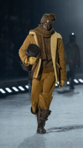 Model walking on a snowy runway at night in a tan shearling coat, brown scarf and knit hat, carrying a rolled hat and gloves.