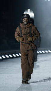 Person in a brown snowsuit and helmet walks toward the camera with a snowboard strapped to their back on a snowy surface filled with haze and lights in the distance.