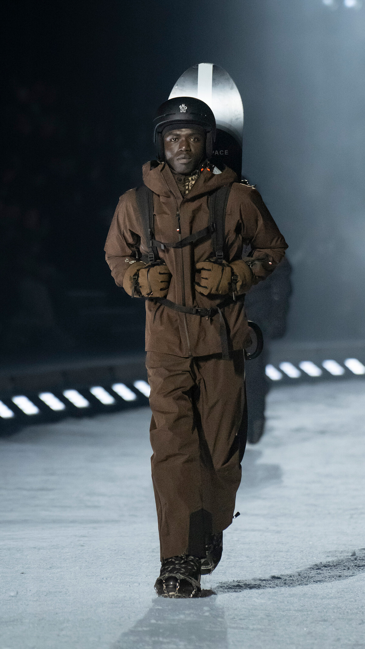 Person in a brown snowsuit and helmet walks toward the camera with a snowboard strapped to their back on a snowy surface filled with haze and lights in the distance.