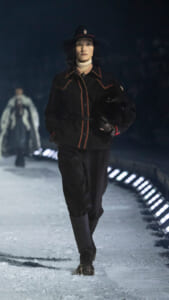 Female model walking a snow-covered runway in a black belted coat with red stitching, wide-brim hat, and gloves.