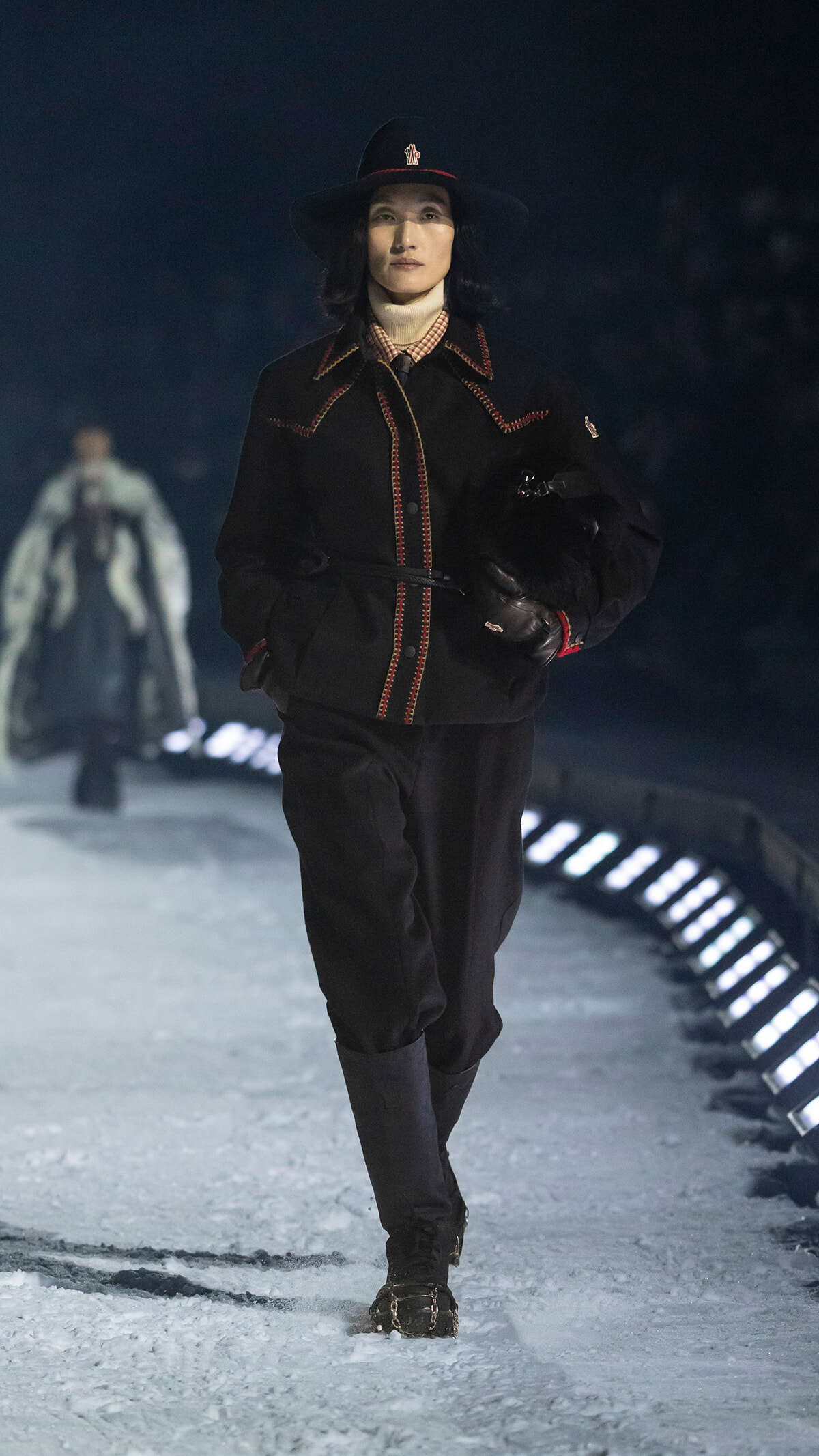 Female model walking a snow-covered runway in a black belted coat with red stitching, wide-brim hat, and gloves.