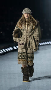Model walks a runway in a chunky Nordic knit coat with matching hat, belt, and fur-trimmed bag on an icy catwalk.
