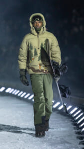 Male snowboarder in a green puffer jacket and goggles, carrying a snowboard, walking on a snowy path at night.