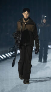 Model walking on a snowy runway at night, carrying a snowboard and wearing a fringed brown‑and‑black jacket with ski goggles on her head.