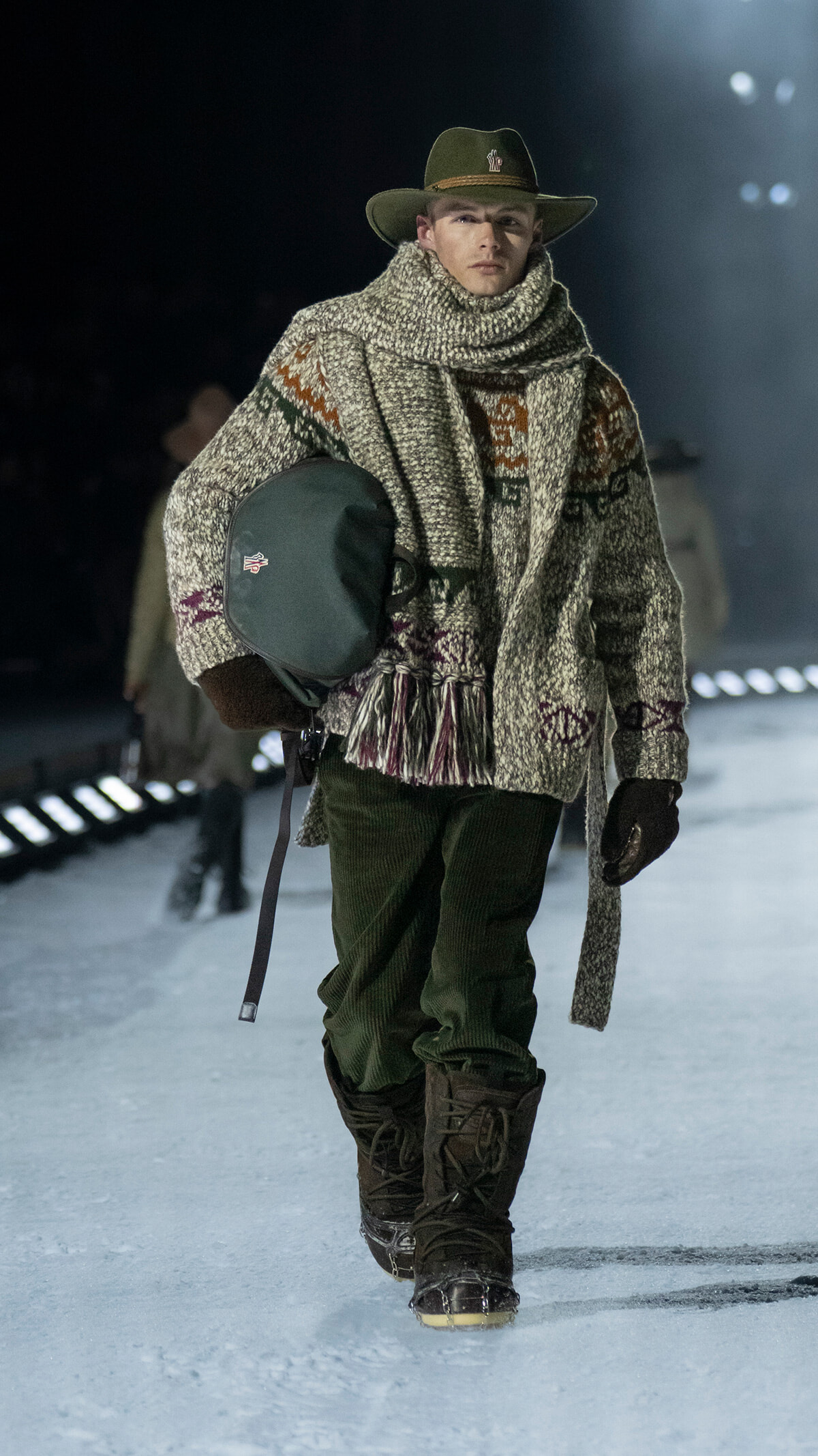 Male model walks on an icy runway in a bulky knit poncho/scarf, green hat, green pants, tall boots, carrying a dark green bag over the shoulder