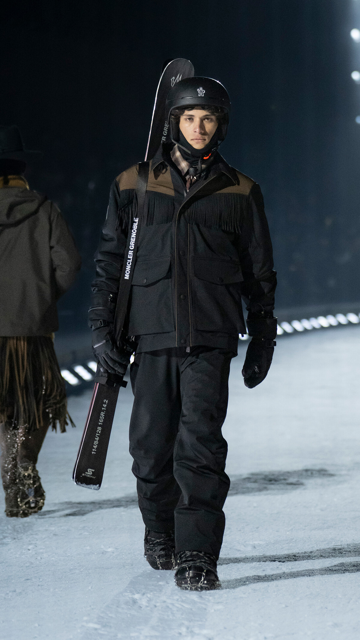 Snowboarder in black winter gear and helmet walking on snow at night, holding a snowboard under his arm.