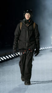 Person in all-black winter gear (jacket, pants, gloves) wearing a helmet and backpack, walking on an icy runway at night.