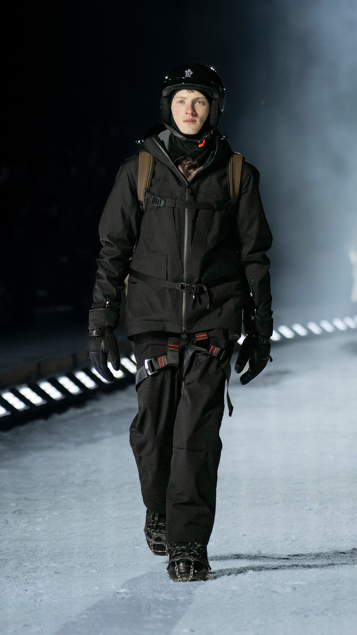 Person in all-black winter gear (jacket, pants, gloves) wearing a helmet and backpack, walking on an icy runway at night.