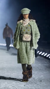 Model walking on a snowy runway in a matching green knit coat and hat, floral-embroidered scarf, and checkered dress with a cream floral bag at the waist; brown boots and dark tights.