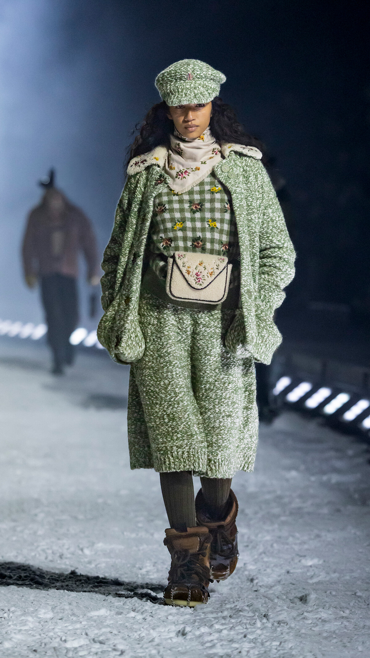 Model walking on a snowy runway in a matching green knit coat and hat, floral-embroidered scarf, and checkered dress with a cream floral bag at the waist; brown boots and dark tights.