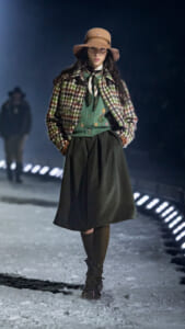 Female model on a snowy runway wearing a beige hat, plaid jacket, green embroidered sweater, and a dark midi skirt.