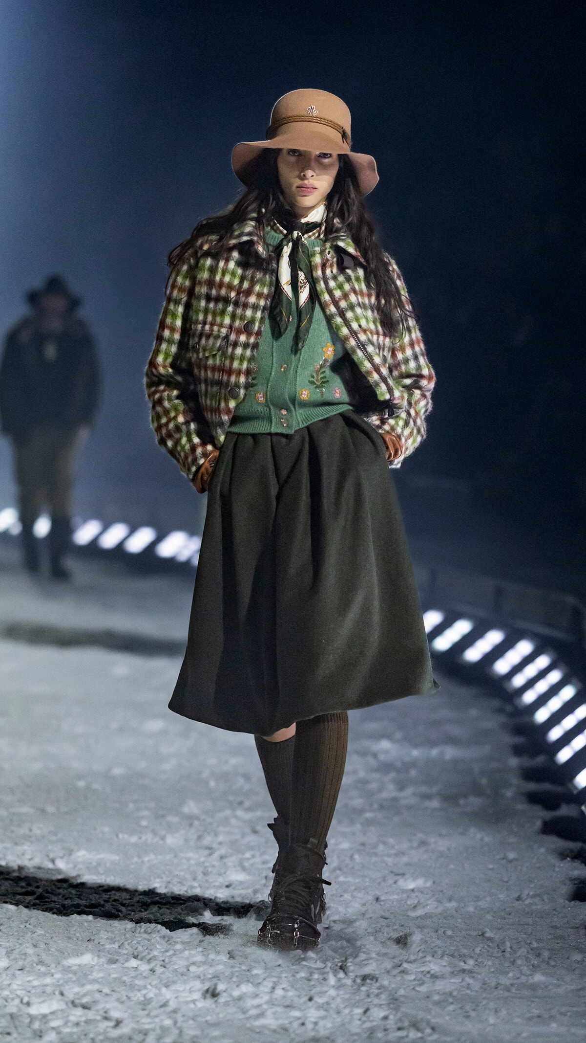 Female model on a snowy runway wearing a beige hat, plaid jacket, green embroidered sweater, and a dark midi skirt.