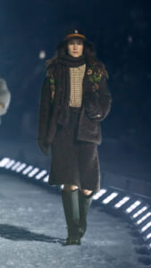 Model walks a snowy runway in a dark fur coat with floral embroidery, checkered shirt, scarf, and tall green boots, wearing a black hat with brown band.