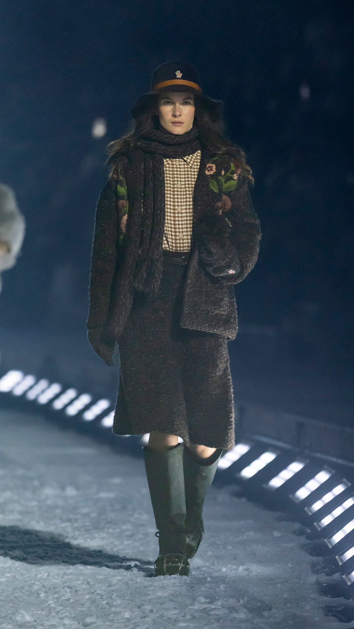 Model walks a snowy runway in a dark fur coat with floral embroidery, checkered shirt, scarf, and tall green boots, wearing a black hat with brown band.