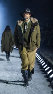 Male model in a green parka with fur trim, scarf, and wide-brim hat walking a snowy, night-time runway.