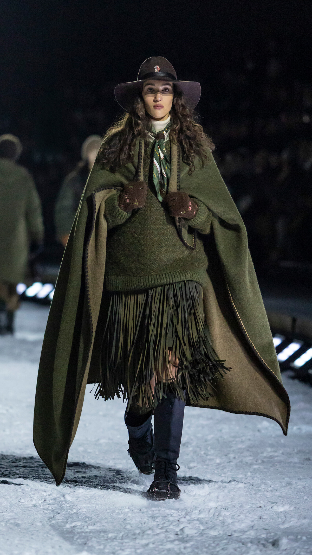 Model walking on a snowy runway at night in an olive-green cape, knit sweater with fringe, gloves, scarf, and a wide-brim hat.