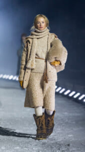 Model walking on a snowy runway in a cream teddy coat, chunky scarf, belt, gloves, and brown lace-up boots, holding a fur accessory under her arm.