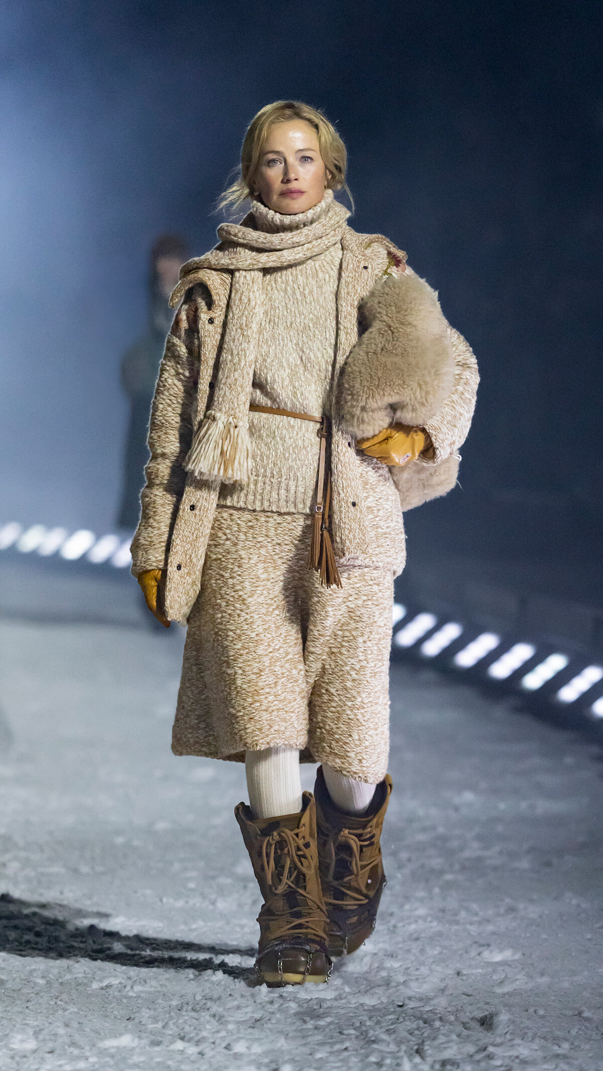 Model walking on a snowy runway in a cream teddy coat, chunky scarf, belt, gloves, and brown lace-up boots, holding a fur accessory under her arm.
