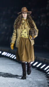 Model walks a snowy runway in vintage brown hat, plaid coat, yellow scarf, and brown skirt, holding a fur muff.