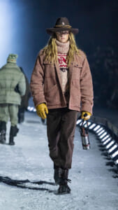Model walking a snowy runway in a brown wide-brim hat, pink tweed jacket, chunky knit sweater, yellow gloves, and a metal water bottle in hand.
