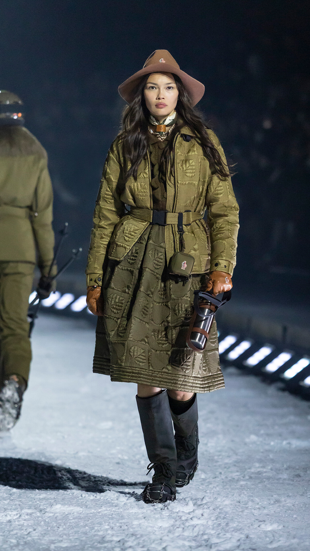 Model wearing olive quilted coat with a belt, brown hat, scarf, leather gloves, and knee-high boots on a snowy runway holding a thermos-like bottle .