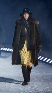 Model walking on a snowy runway in a long brown fur coat, floral dress, black hat, and gloves for a fashion show.