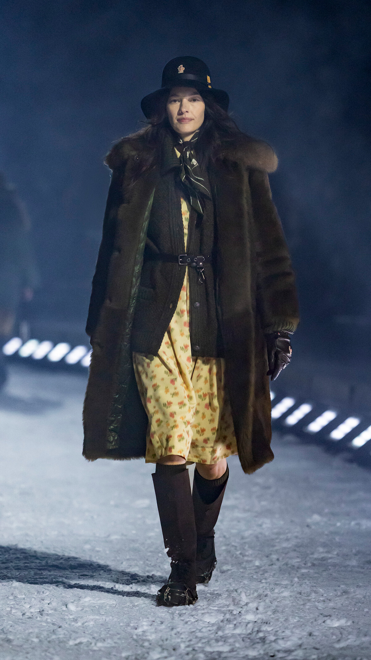 Model walking on a snowy runway in a long brown fur coat, floral dress, black hat, and gloves for a fashion show.