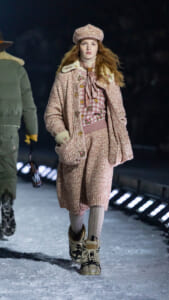 Model on an icy runway wearing a pink fuzzy coat and matching hat, floral pink sweater, scarf, and chunky boots, with knee-high socks against a dark backdrop.