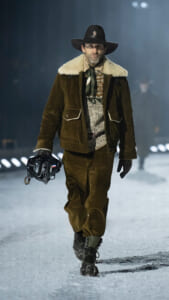 Male model walking a snowy runway in a brown corduroy jacket with shearling collar, scarf, and wide-brim hat, holding a black ski helmet in one hand.