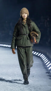 Woman walking in a green quilted winter coat, brown beanie, and gloves on a snowy path at night.