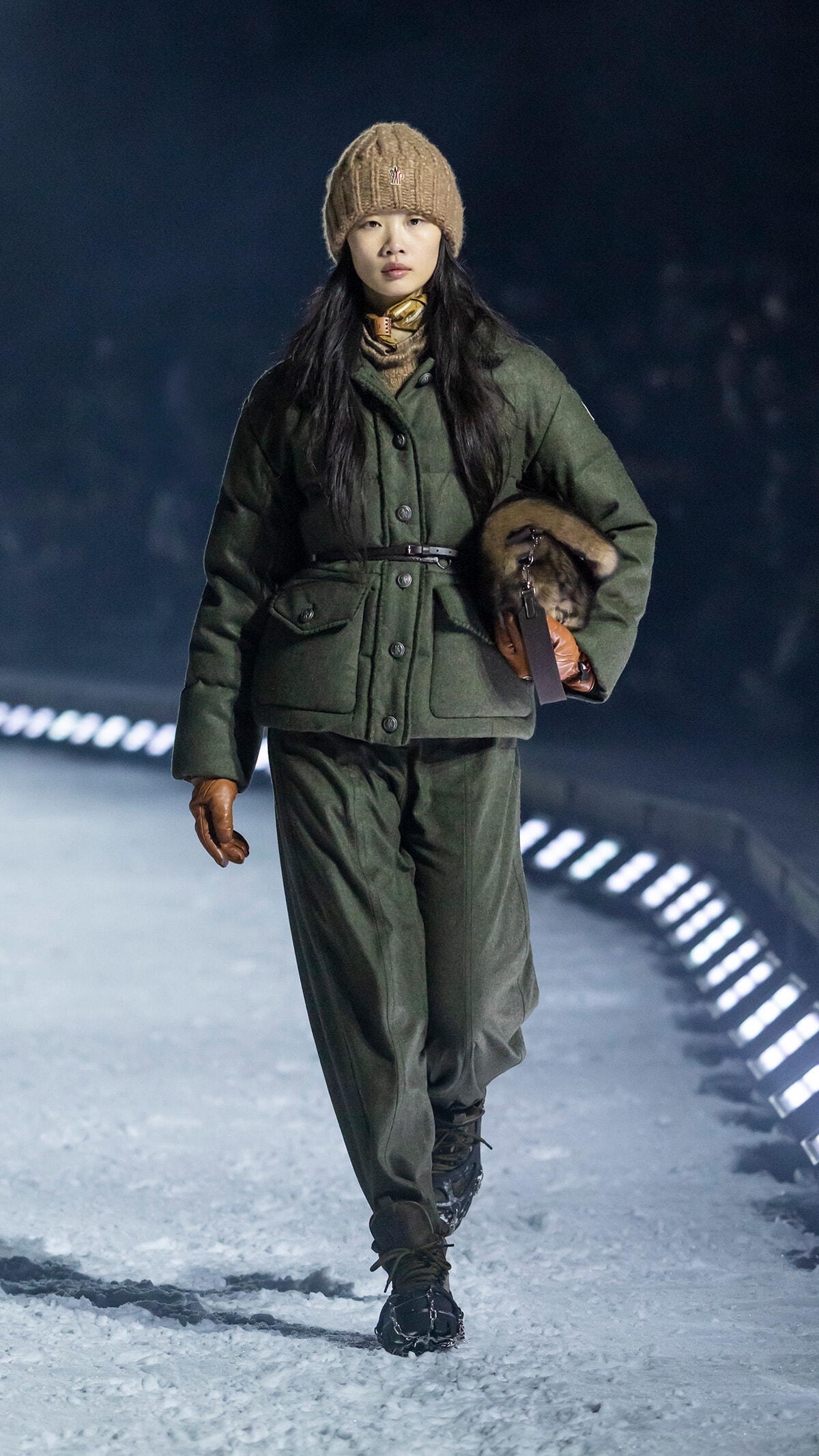 Woman walking in a green quilted winter coat, brown beanie, and gloves on a snowy path at night.