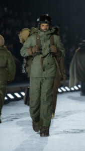 Person in green winter parka and pants, wearing a helmet and goggles, carrying a large pack, walking on an icy surface at night.