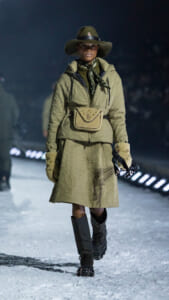 Model walking a snowy runway in an olive quilted coat, wide-brim hat, scarf, beige belt bag, and fur-lined gloves with tall black boots
