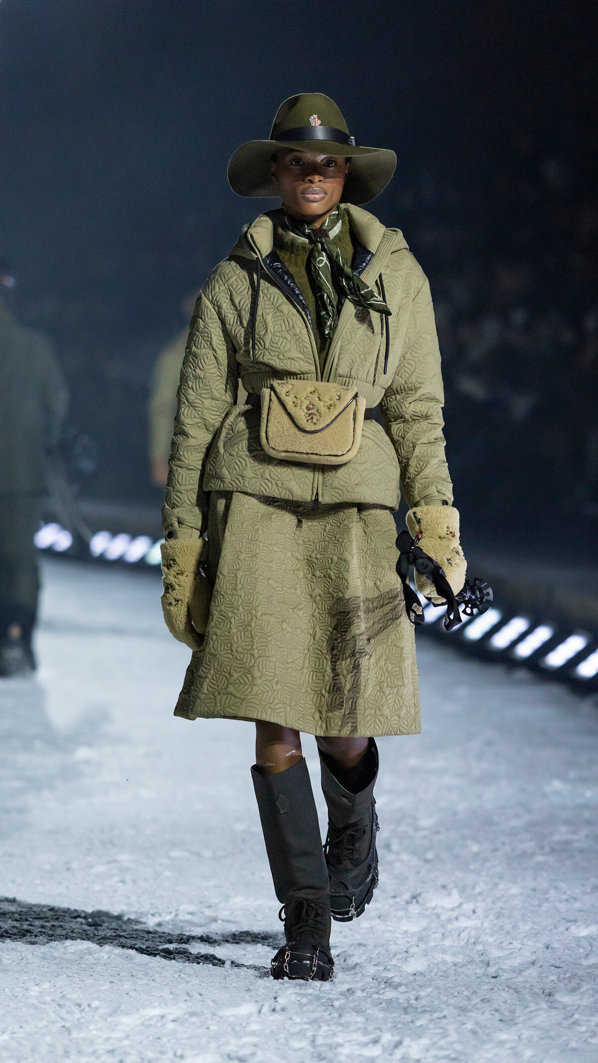 Model walking a snowy runway in an olive quilted coat, wide-brim hat, scarf, beige belt bag, and fur-lined gloves with tall black boots