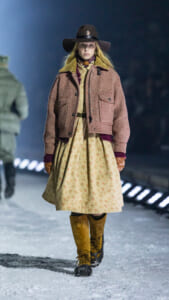 Model walking a snowy runway in a vintage-inspired outfit: floral dress under a brown tweed jacket, mustard knee-high boots, wide-brimmed hat.