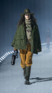Female model walking a snowy runway in an olive-green parka, chunky knit turtleneck, brown pants, and tall green boots, wearing a wide-brimmed hat and carrying a lantern-style bottle by a leather strap in one hand.