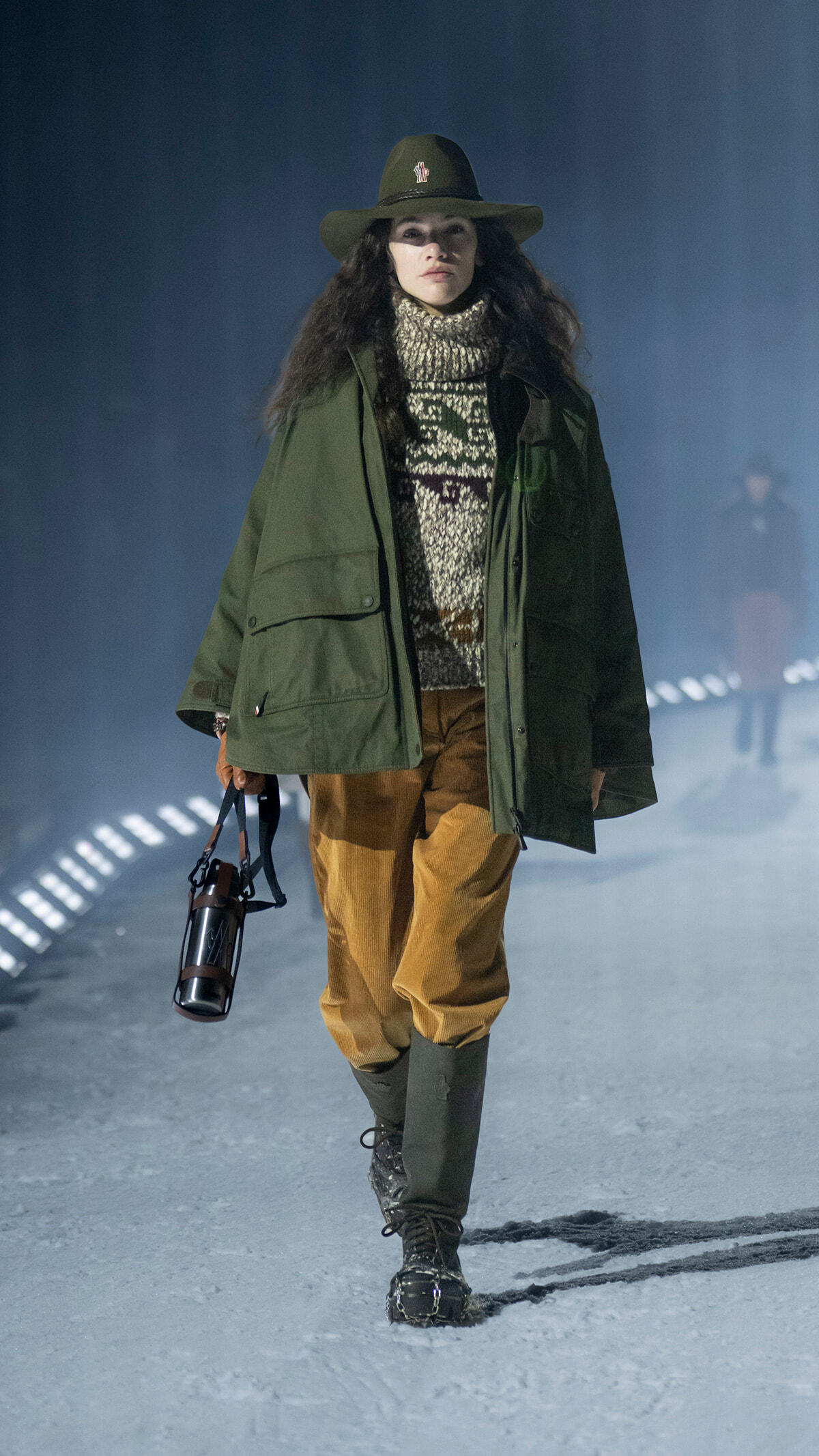 Female model walking a snowy runway in an olive-green parka, chunky knit turtleneck, brown pants, and tall green boots, wearing a wide-brimmed hat and carrying a lantern-style bottle by a leather strap in one hand.