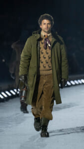 Male model walking a snowy runway in a green parka, beige reindeer-patterned sweater, corduroy pants, boots, and a metal thermos in hand at night fashion show, conveying a rugged winter look.