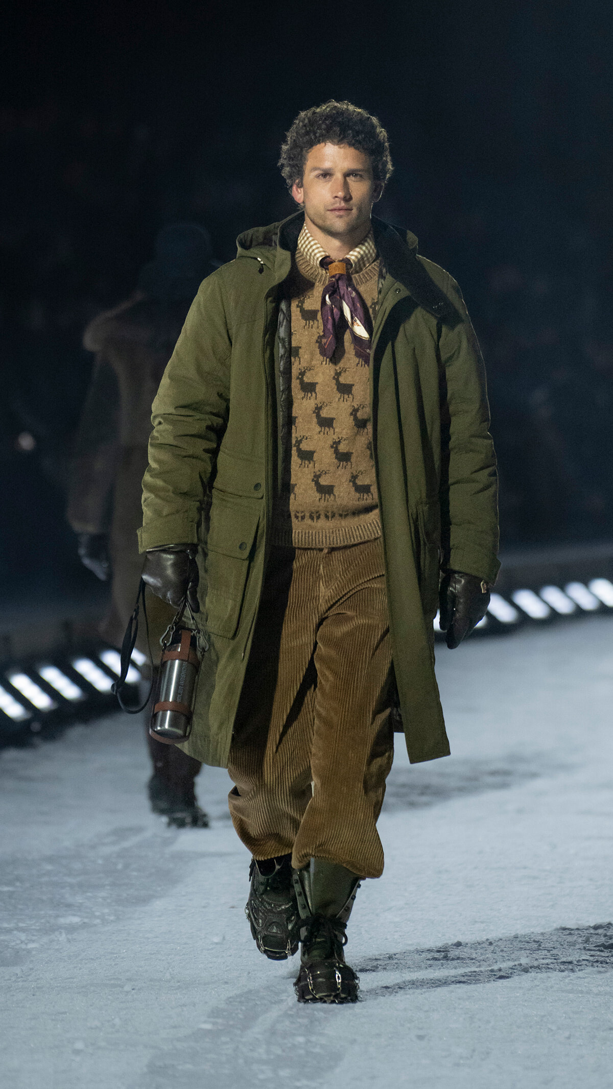 Male model walking a snowy runway in a green parka, beige reindeer-patterned sweater, corduroy pants, boots, and a metal thermos in hand at night fashion show, conveying a rugged winter look.