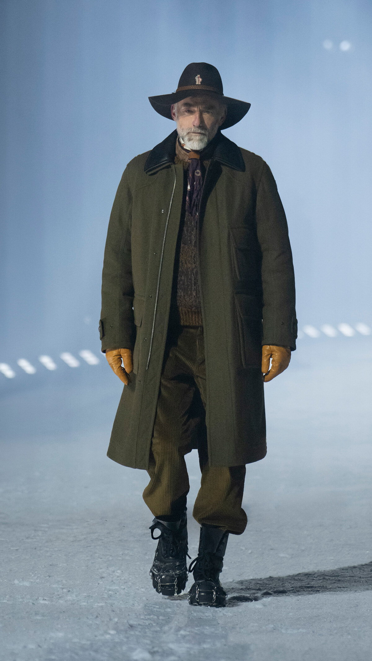 Man in a long olive-green overcoat and brown gloves walks on a snowy surface, wearing a wide-brimmed black hat.