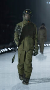 Model wearing olive green winter gear and helmet, carrying a snowboard, walking a snowy runway during a fashion show and looking ahead.