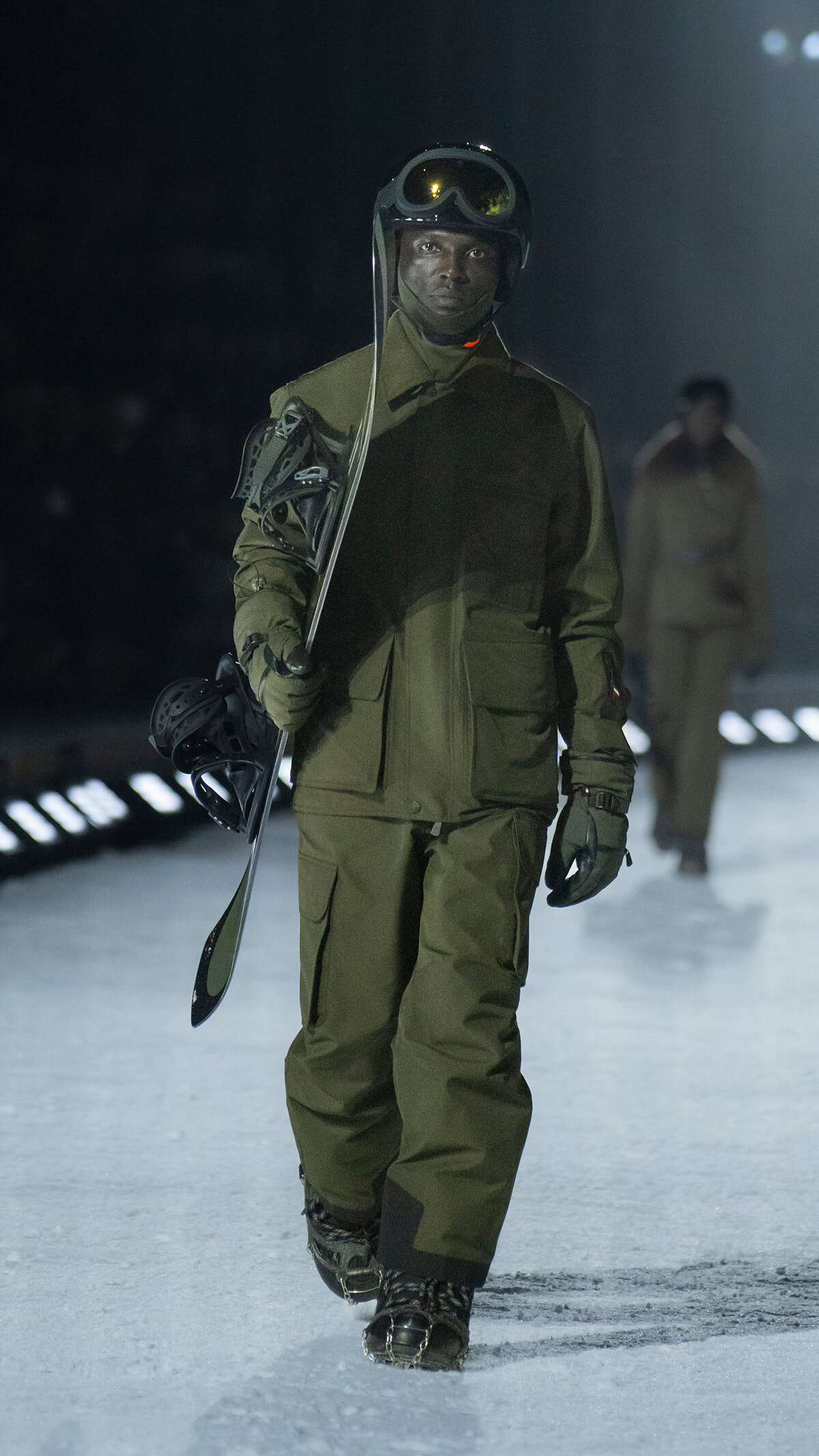 Model wearing olive green winter gear and helmet, carrying a snowboard, walking a snowy runway during a fashion show and looking ahead.