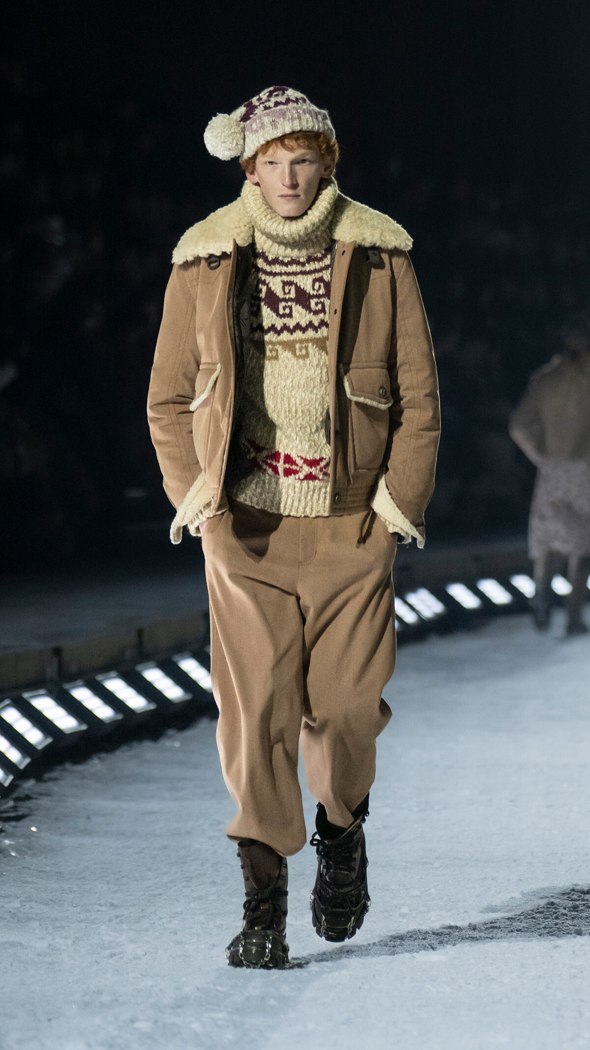 Male model walking a snowy runway in a beige shearling-lined tan coat, cream knit sweater, and matching hat with boots.
