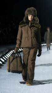 Model walking on a snowy runway in a brown fur-trimmed parka and hat, carrying a large tan duffel bag.