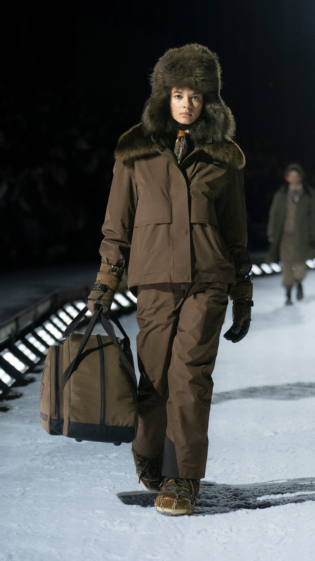 Model walking on a snowy runway in a brown fur-trimmed parka and hat, carrying a large tan duffel bag.